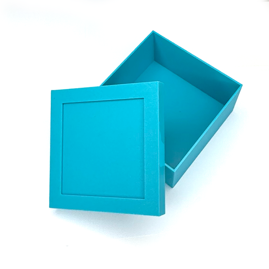 A bright turquoise blue square cardboard box with its lid leaning against the front side, showing the inside of the box and a recessed square panel on the lid, set against a plain white background.